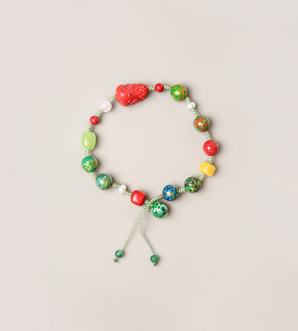 A beaded stretch bracelet with a mix of green, red, yellow, and blue marbled beads, a textured red heart-shaped bead, and a light green cord with sliding beads, displayed on a light beige background.