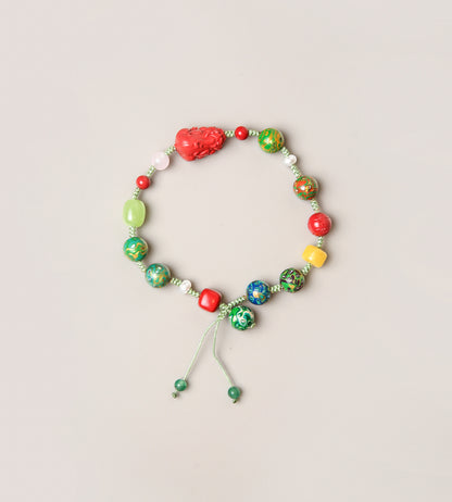 A beaded stretch bracelet with a mix of green, red, yellow, and blue marbled beads, a textured red heart-shaped bead, and a light green cord with sliding beads, displayed on a light beige background.