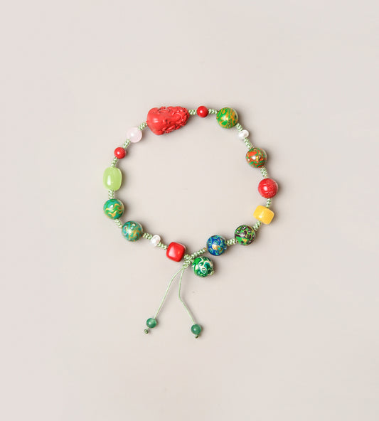 A beaded stretch bracelet with a mix of green, red, yellow, and blue marbled beads, a textured red heart-shaped bead, and a light green cord with sliding beads, displayed on a light beige background.